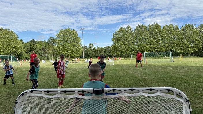 Kinder und ein Trainer spielen auf einem Sportplatz Fußball. Im Vordergrund steht ein Junge in einem Fußballtor.  