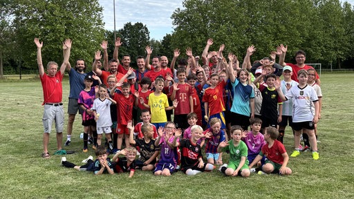rund 60 Menschen in Sportkleidung stehen auf einem Sportplatz und winken fröhlich in die Kamera