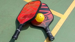 Zwei Pickleball-Schläger und ein Ball liegen auf einem Feld.