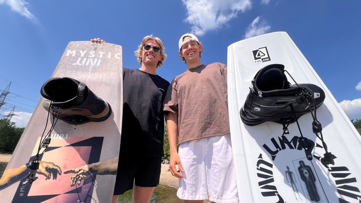 Zwei junge Sportler stehen mit ihren Wakeboards vor einem See