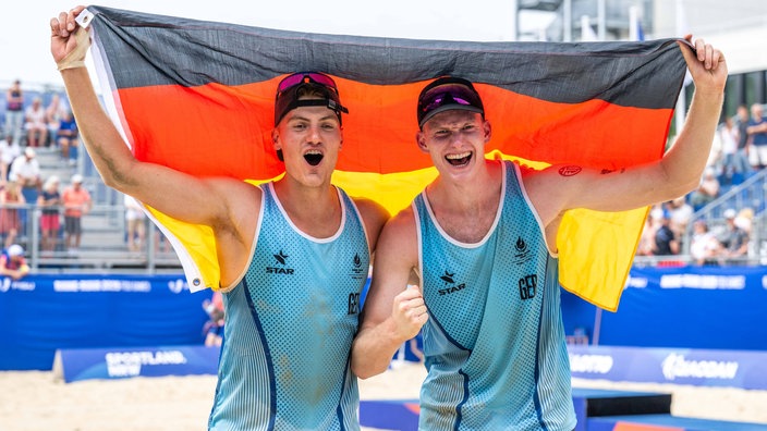 Maximilian Just und Philipp Huster jubeln mit einer Deutschland-Fahne auf dem Beachvolelyball-Court.