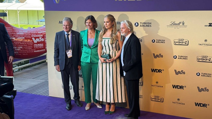 Stefanie Peters im grünen Anzug und Prinzessin Amalia im weißen Kleid mit grünen Streifen, flankiert von zwei Männern im Anzug.