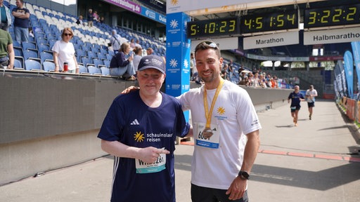 Zwei Männer stehen gemeinsam in einem Marathon-Zieleinlauf und lächeln in die Kamera. Einer von ihnen ist der Ausdauersportler Joey Kelly.
