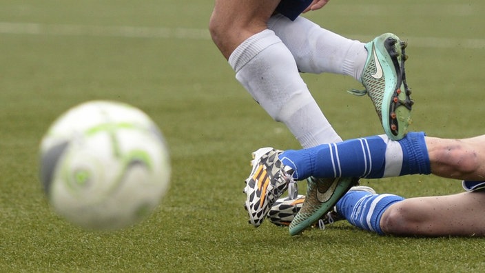 Zu sehen sind die Beine zweier Fußballspieler. Einer grätscht den anderen von hinten ab. Im Vordergrund ein Ball.  