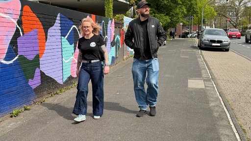 Eine blonde Frau mit Brille und ein Mann mit Brille, Bart und Kappe laufen auf einem Bürgersteig entlang