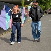 Eine blonde Frau mit Brille und ein Mann mit Brille, Bart und Kappe laufen auf einem Bürgersteig entlang