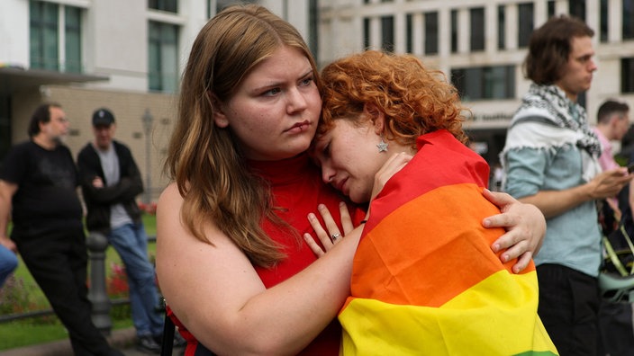 Eine Frau in einer Regenbogenfahne gehüllt lehnt weinend an der Schulter einer weiteren Frau