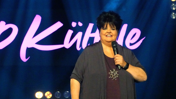 Einfe Frau auf der Bühne. Sie hält ein Mikro in der Hand, hinter ihr sieht man einen Schriftzug "Kühne"