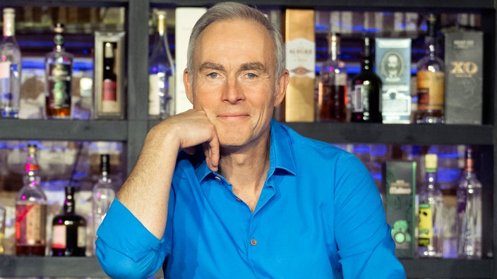 Johann König schaut in die Kamera. Dahinter eine Bar mit Getränkeauswahl