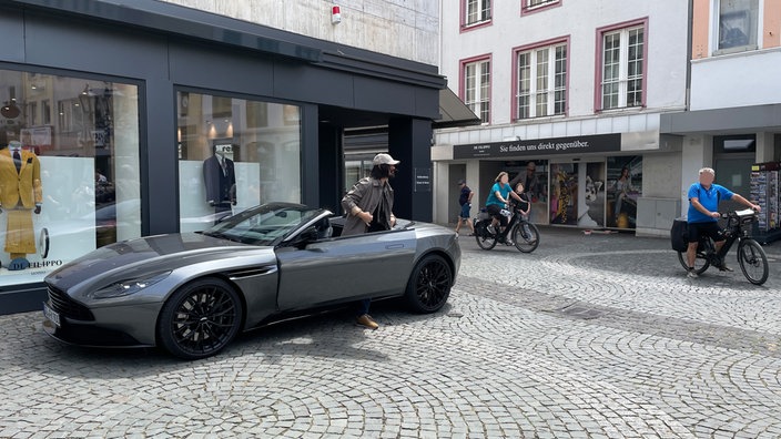 Ein silberner Aston Martin vor einem Geschäft, ein Mann mit Kappe steigt aus