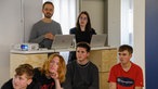 Eine Gruppe Jugendliche und zwei Erwachsene schauen einer Präsentation zu