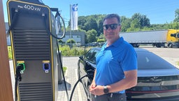 Mann mit Sonnenbrille steht vor seinem E-Auto an einer Ladesäule