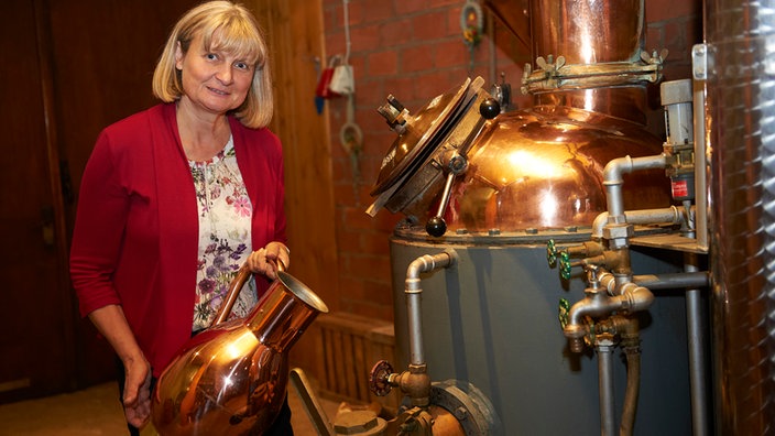  Margit Paschen in der Schnapsbrennerei, sie steht mit einem Kupferkrug vor dem Brennkessel
