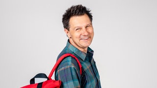André Gatzke steht mit dem roten Wunderschön-Rucksack vor einer weißen Wand