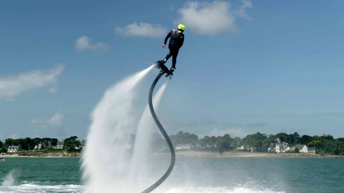 Ein Mann mit Helm steht auf einem Brett und wird von einem Schlauch mit Wasserkraft hoch geschleudert