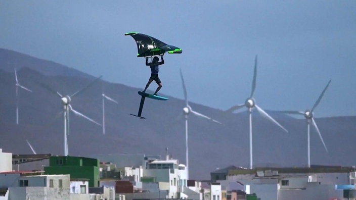 Ein Surfer mit einem Drachen fliegt durch die Luft, im Hintergrund Windkraftanlagen