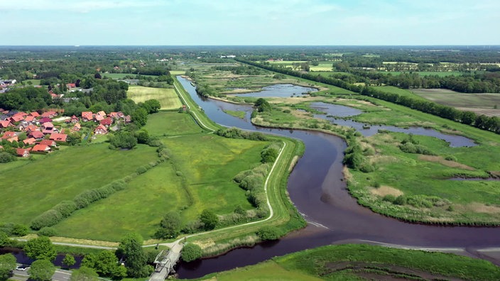 Blick von oben auf eine Wiesen-Wasser-Landschaft mit einem kleinen Dorf