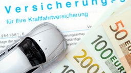 Das Bild zeigt einen Versicherungsvertrag, auf dem einige Geldscheine und ein silbernes Spielzeugauto liegen