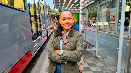Eine junge Frau steht an einer Straßenbahnhaltestelle in Düsseldorf und lächelt in die Kamera
