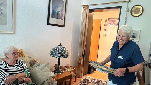 Monika Gomulia hilft einer anderen Rentnerin im Haushalt. Während Hedwig Anna Nitsch auf der Couch sitzt, läuft Monika Gomulia mit einem Stapel Wäsche durch die Wohnung.