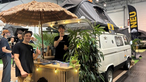 Zwei junge Männer stehen unter einem Strohschirm an einem Stand vor einem VW Bully mit Dachzelt