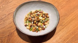 Ein Teller mit „Blumenkohl-Ceviche mit Rauchmandeln“ steht auf einem Holztisch.