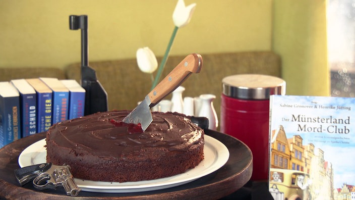 Schokoladenkuchen auf einem Drehteller, links daneben liegt eine kleine Pistole. Mitten in dem Kuchen ragt ein großes Küchenmesser heraus, an dem Marmelade klebt. Rechts daneben ein Buch mit dem Titel: Der Münsterland Mord-Club.