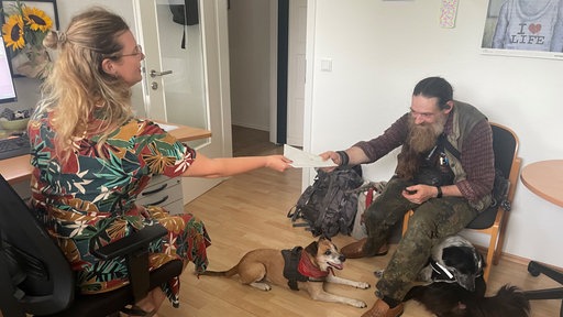 Ein älterer Mann mit langem Baart sitzt auf einem Stuhl und überreicht einer Frau Briefe.