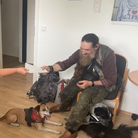 Ein älterer Mann mit langem Baart sitzt auf einem Stuhl und überreicht einer Frau Briefe.