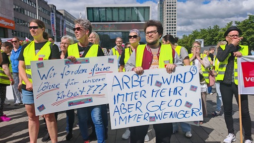 Streikende mit Schildern in den Händen mit dem Aufdruck "Wir fordern faire Löhne für unsere Arbeit!!!" und "Arbeit wird immer mehr, aber Geld immer weniger!"