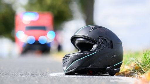 An einem Straßenrand liegt ein Motorradhelm. Im Hintergrund ist unscharf ein Krankenwagen.