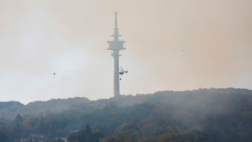 Helikopter mit Löschwasser-Außenlastbehälter beim Waldbrand im Hürtgenwald vor einem Turm