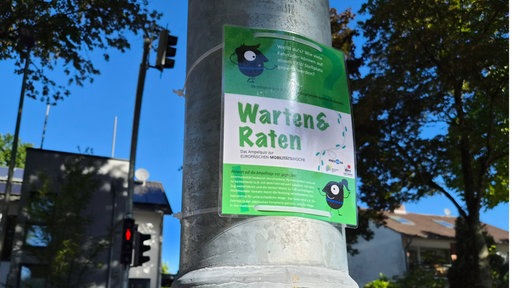 Ein grüner Zettel mit dem Warten-und-Raten-Logo hängt an dem Pfosten einer Ampel.