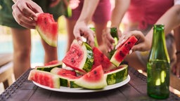 Auf einem Tisch steht ein Teller mit geschnittener Wassermelone, mehrere Personen greifen zu