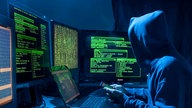 Eine Person sitzt im Dunkeln mit Smartphone vor mehreren Computern
