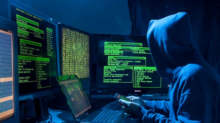 Eine Person sitzt im Dunkeln mit Smartphone vor mehreren Computern