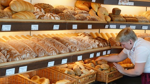 Eine Verkäuferin steht in einer Bäckerei vor einem Regal mit Brötchen und Broten.