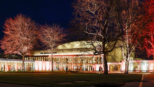 Blick auf die gelb-rot erleuchtete Beehovenhalle in Bonn, umgeben von ein paar Bäumen, die rot angestrahlt werden
