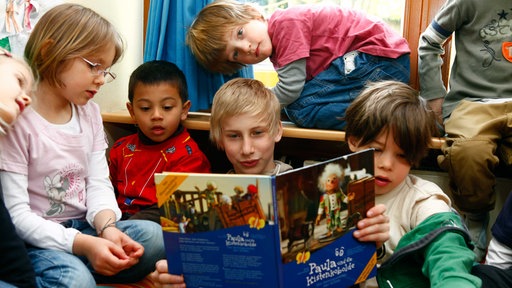 Ein junge mit blonden Haaren sitzt auf dem Boden und hat ein Buch aufgeschlagen und liest daraus vor, um ihn herum sitzen jüngere Kinder