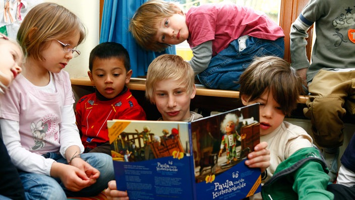 Ein junge mit blonden Haaren sitzt auf dem Boden und hat ein Buch aufgeschlagen und liest daraus vor, um ihn herum sitzen jüngere Kinder
