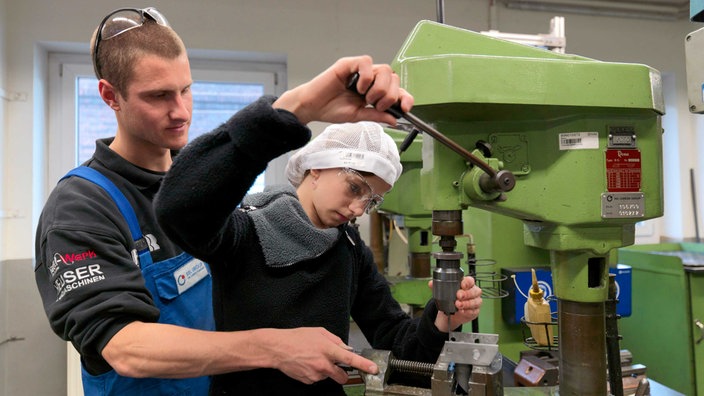 Ein Mädchen mit schwarzem Pulli und weißter Haube steht an einer grpnen Werkbank, hinter ihr steht ein Mann im Blaumann 