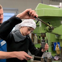 Ein Mädchen mit schwarzem Pulli und weißter Haube steht an einer grpnen Werkbank, hinter ihr steht ein Mann im Blaumann 