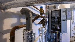 Eine Gasbrennwerttherme hängt im Heizkeller eines Mehrfamilienhauses. Neben dem geöffneten Gerät sind Rohrleitungen, Abgasführung und Armaturen der Heizungsanlage zu sehen.