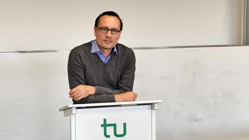 Ein Mann mit einem grauen Pullover und einem blauen Hemd darunter, der auf einem Stehpult mit einem grünen "tu" darauf vor einem Whitboard lehnt.