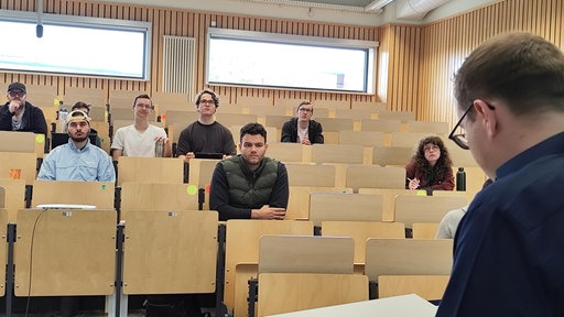 Der Rücken eines Mannes mit Brille, der an einem Pult steht, davor junge Menschen, die in hörsaaltypischen Reihen sitzen.