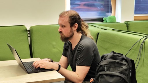 Ein Mann mit langen Haaren, der auf einem grünen Sofa an einem Laptop sitzt.