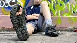 Ein Junge sitzt vor einer Wand und streckt seinen kaputten Turnschuh mit durchgelaufener, löchriger Sohle in die Kamera.