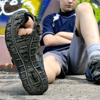 Ein Junge sitzt vor einer Wand und streckt seinen kaputten Turnschuh mit durchgelaufener, löchriger Sohle in die Kamera.