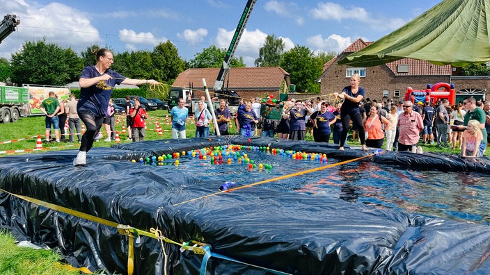 Zwei Frauen stehen auf einem mit Silofolie ausgelegten Spielfeld, in dem bunte Plastikbälle liegen und balancieren durch Seile einen grünen Korb, der mit bunten Bällen gefüllt ist