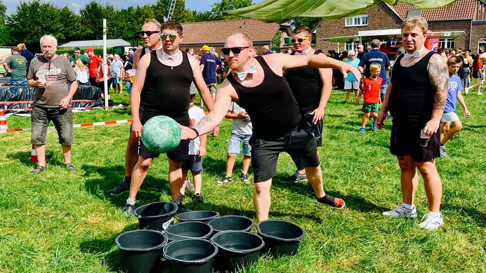 Fünf Männer in schwarzen Tanktops und weißer Fliege stehen vor acht angereihten Eimern. Einer von ihnen hat einen Fußballgroßen Ball in der Hand und setzt zum Wurf an